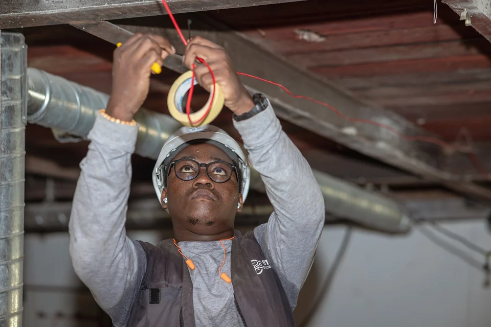 An electrician in a white hard hat and glasses reaches overhead with both hands, holding a roll of tape and a red wire under a ceiling with metal ducting