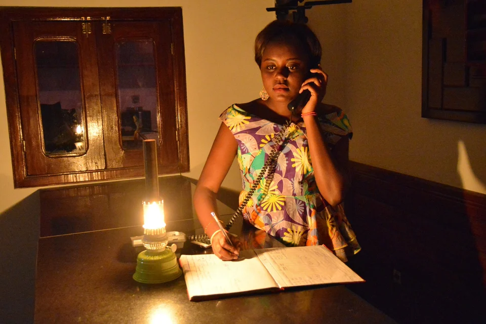 A hotel receptionist holds a corded phone to her ear and writes in an open paper ledger by the light of an oil lamp at a dark front desk