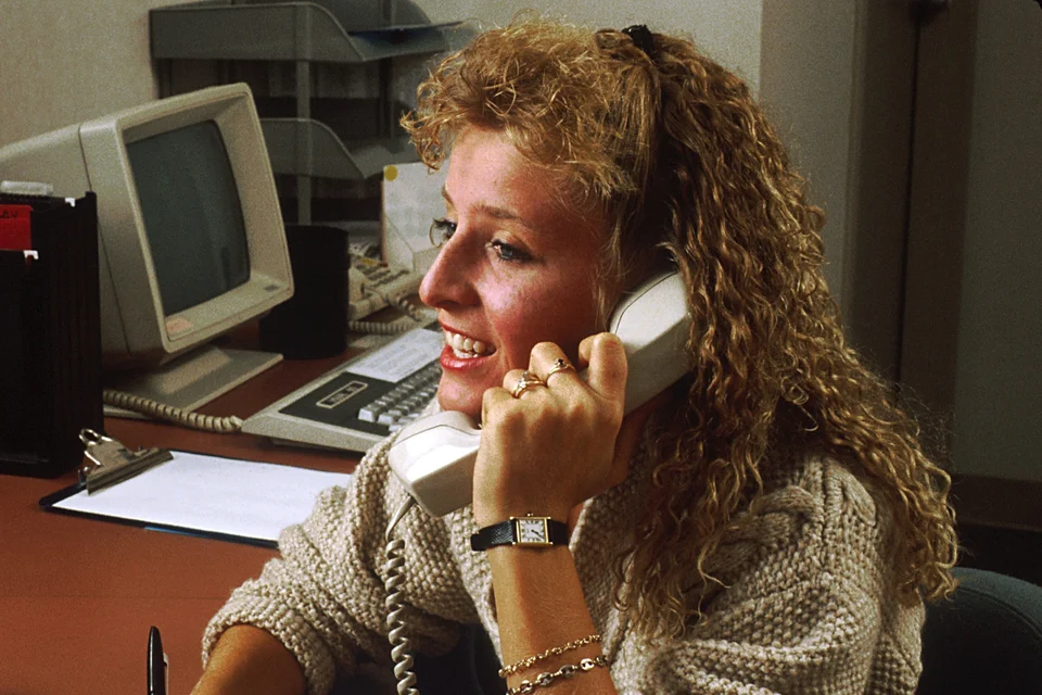 A receptionist at an office desk smiles while speaking on a corded telephone, with a computer monitor and office equipment on the desk