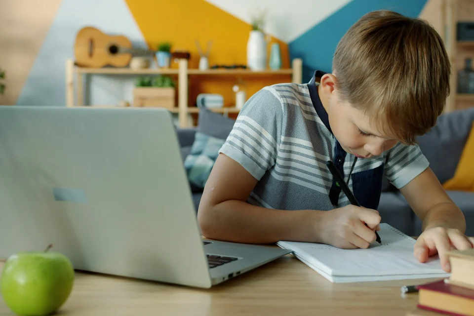 A child writing in a notebook beside a laptop