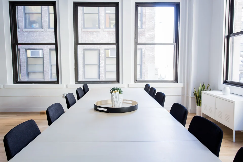 A long white meeting table with a row of empty black chairs along both sides in front of tall windows