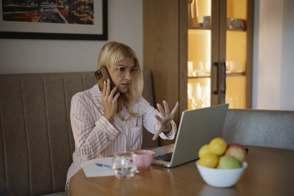 A woman in glasses holds a smartphone to her ear and gestures at an open laptop while sitting at a table at home